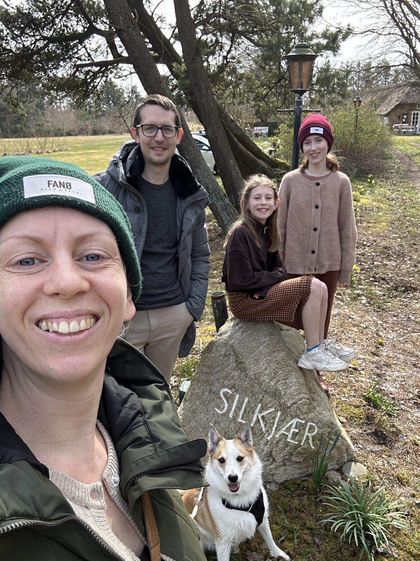 Familie med hund foran en stor kampesten med navnet SILKJÆR hugget ind, den stråtækte gård i baggrunden