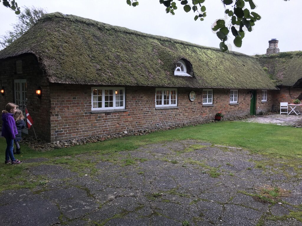 Den lange stråtækte længe med hvide vinduer, kampestenssokkel og to børn med Dannebrog
