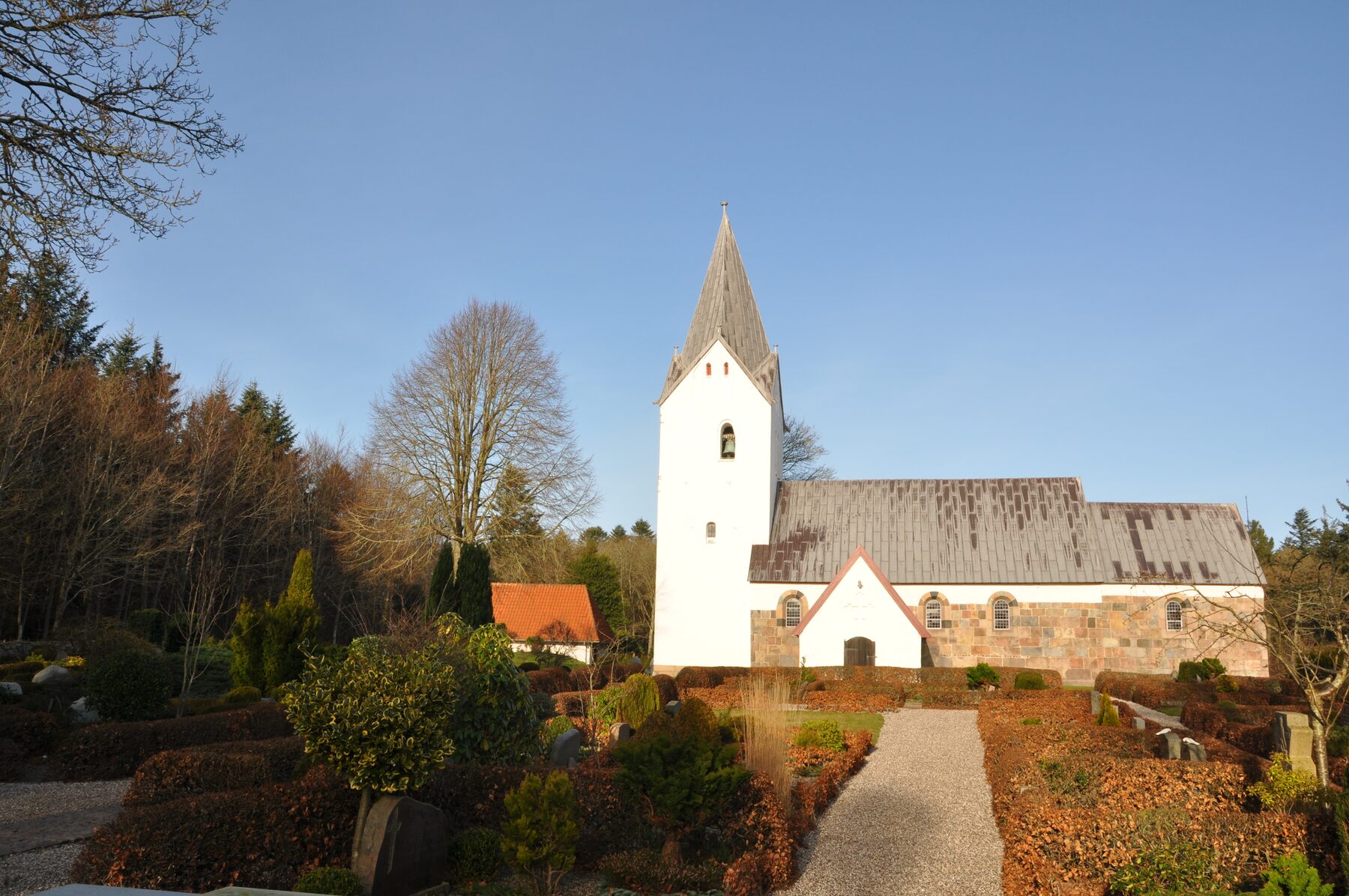 Husby Kirke: hvidkalket tårn med spir, granitkvadre i skibets murværk og kirkegård med klippede bøgehegn og gravsten