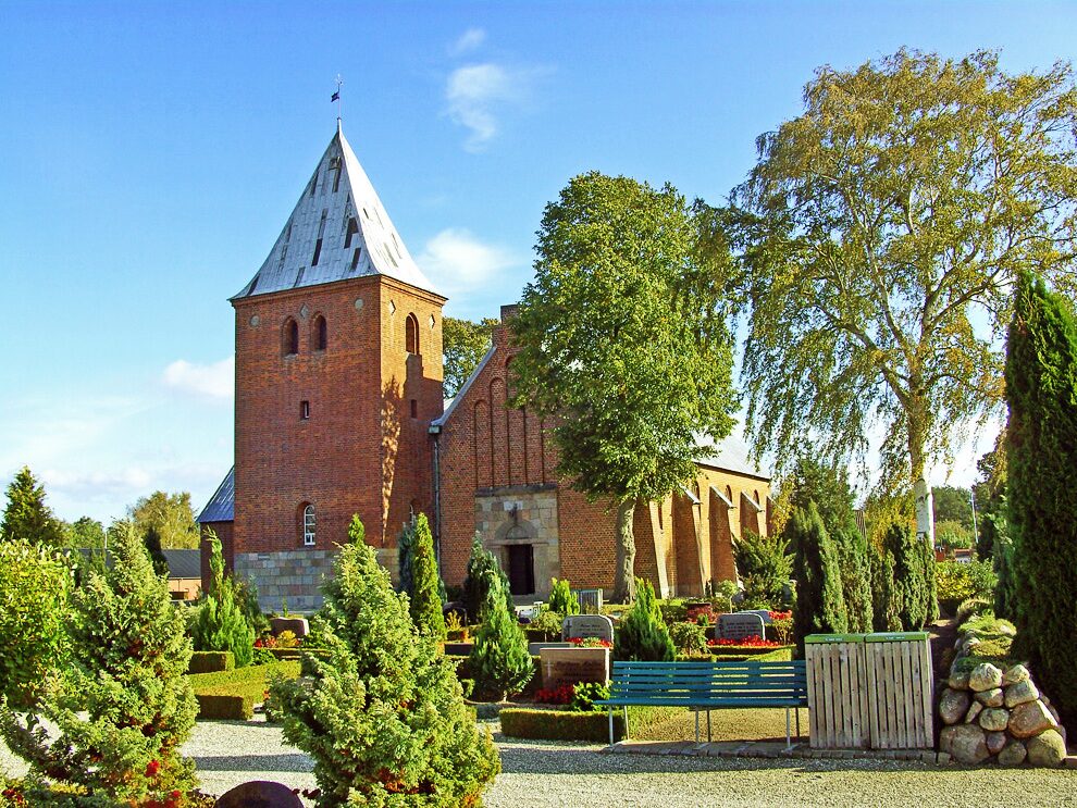 Vejlby Kirke ved Allingåbro: tårn af røde munkesten med blytag, granitkvadre nederst i murværket, kirkegård med thujaer og gravsteder
