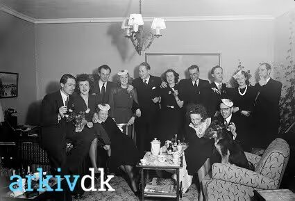 Selskab i en dagligstue omkring 1944: fjorten voksne opstillet foran sofa og rullebord med kaffestel, nogle med små papirhatte på hovedet