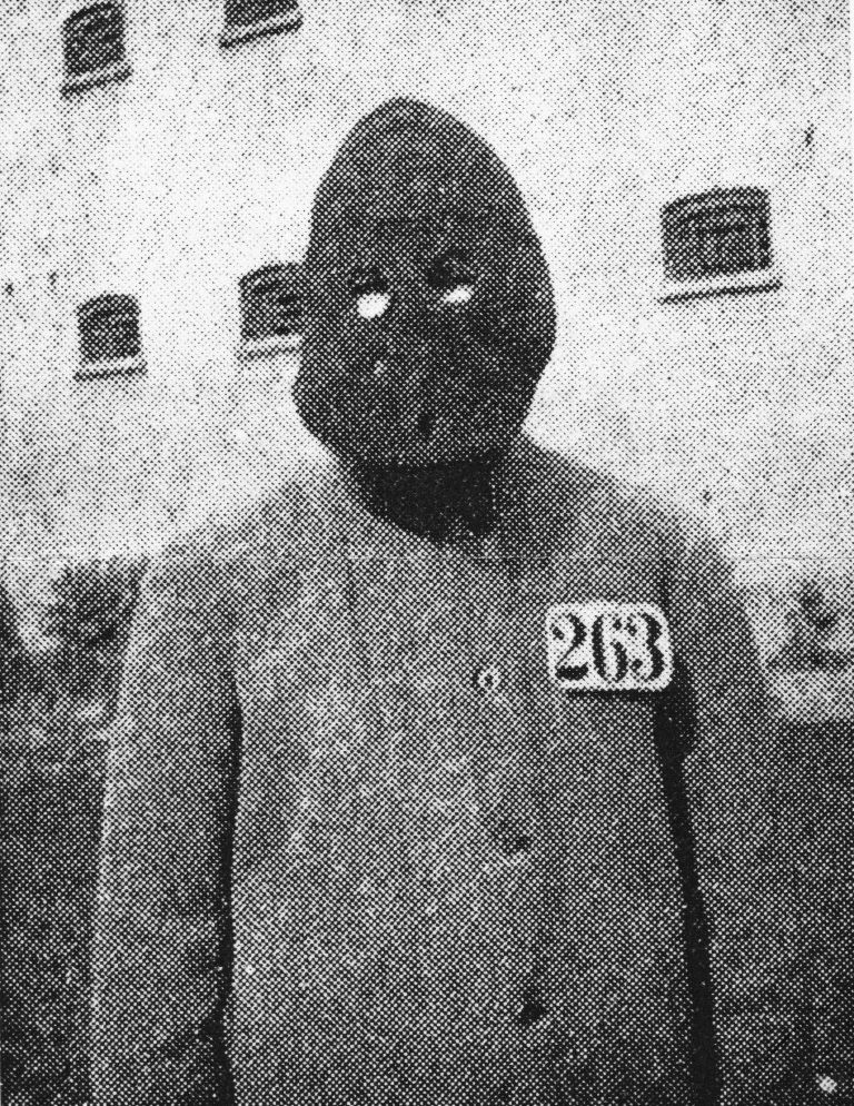 Fange i Vridsløselille Straffeanstalt fotograferet foran cellebygningen, iført anstaltens dragt med fangenummer 263 på brystet og Philadelphia-systemets maske, der dækker hele hovedet med huller til øjnene