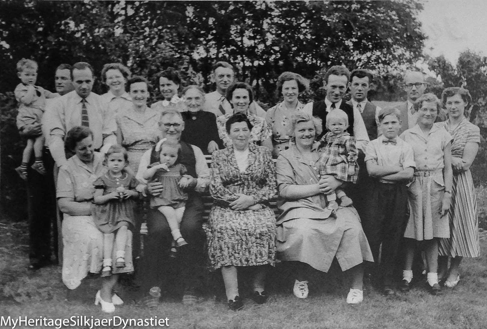 Sort-hvid gruppefoto af omkring femogtyve mennesker i en have, tre generationer, opstillet omkring en bænk med et ældre par siddende i midten